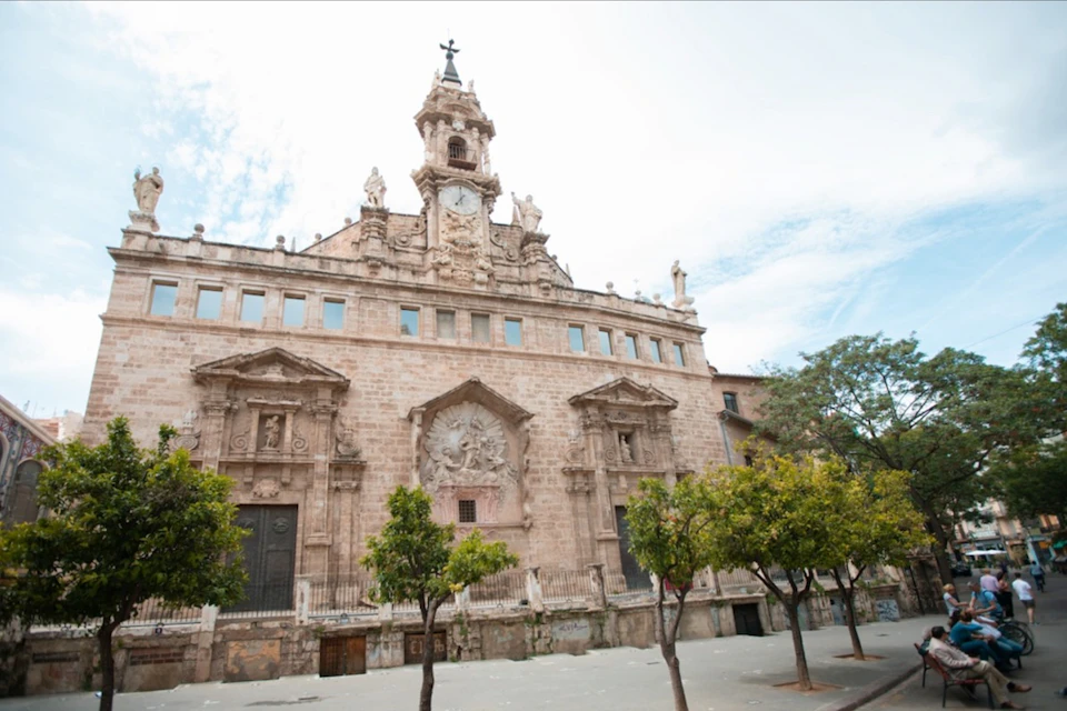 Historic stone building with ornate architectural details, large clock tower, and trees in the foreground under a partly cloudy sky.