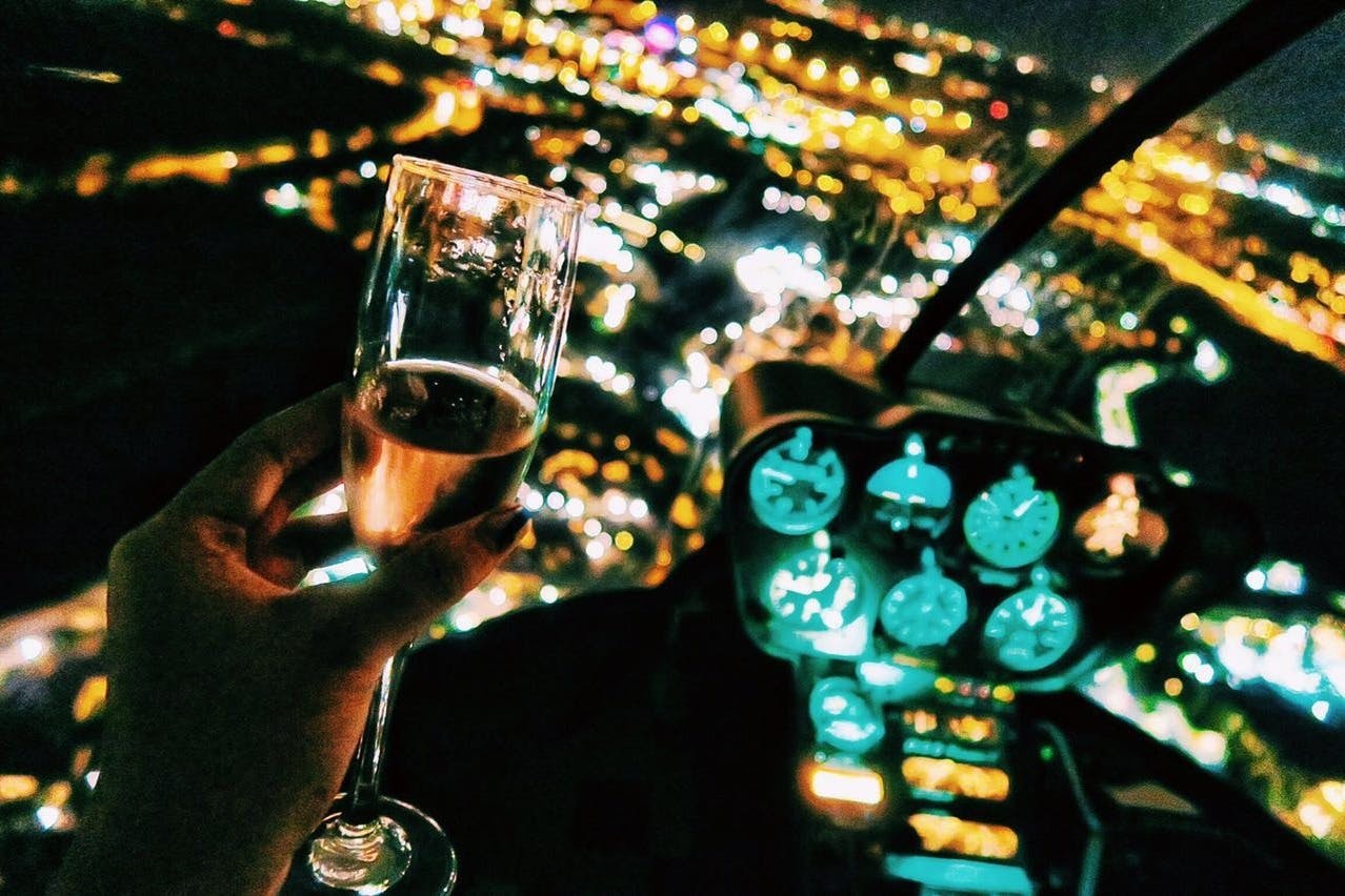 A hand holds a champagne glass inside a helicopter cockpit, with a nighttime cityscape illuminated by lights in the background.