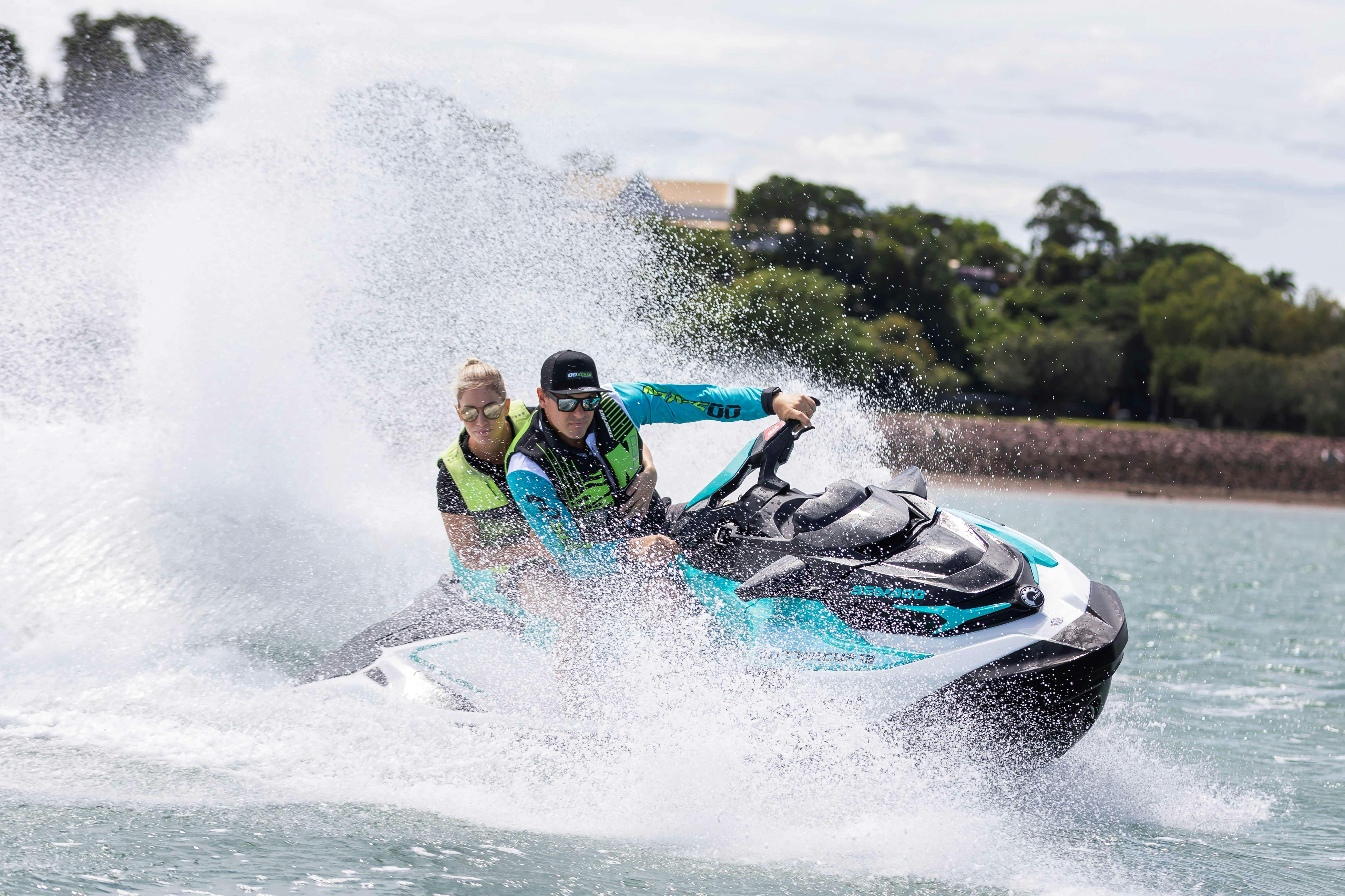 Δύο άνθρωποι σε ένα τζετ σκι δημιουργούν μια μεγάλη βουτιά ενώ επιταχύνουν μέσα στο νερό, φορώντας σωσίβια και γυαλιά ηλίου.