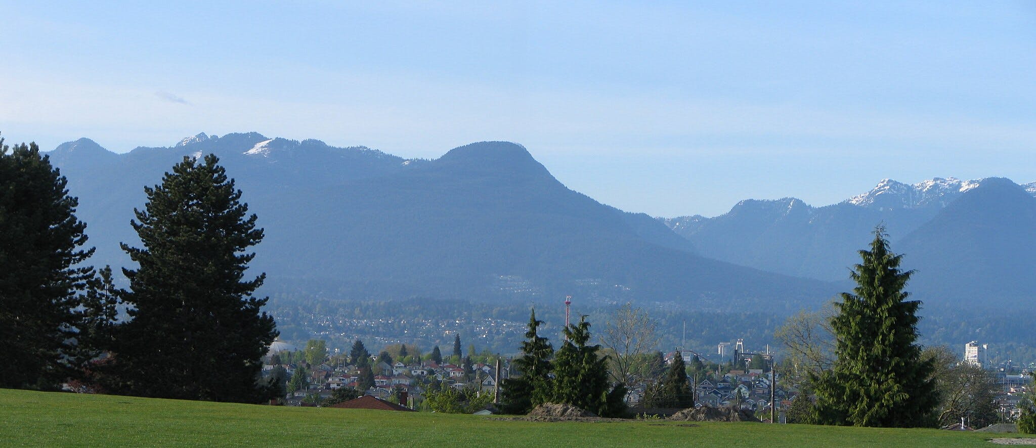 Montagnes enneigées sous un ciel bleu avec un paysage urbain et de la verdure au premier plan.