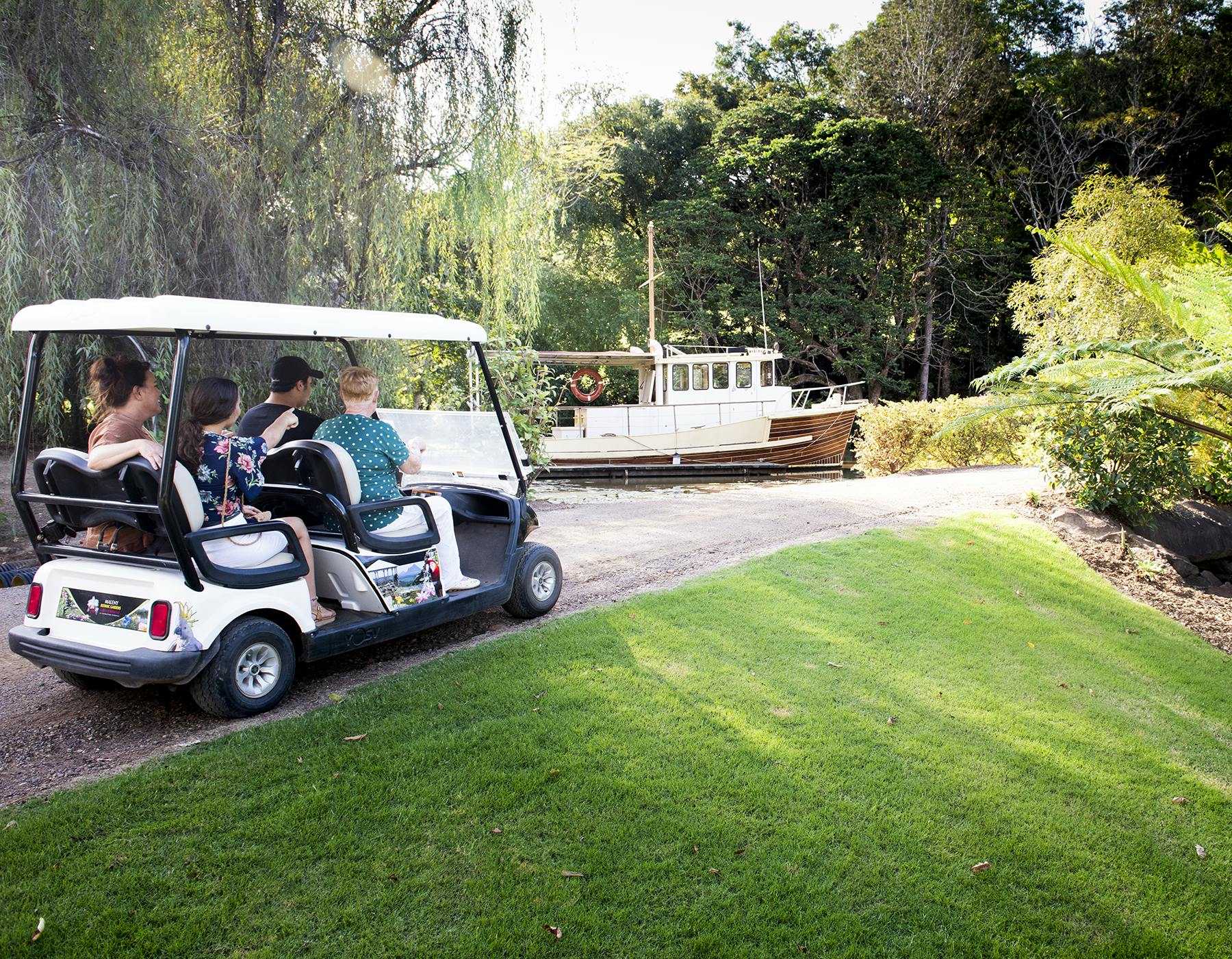 Niewielka grupa ludzi jedzie wózkiem golfowym ścieżką w pobliżu bujnego zielonego trawnika, z białą łodzią i gęstymi drzewami w tle.