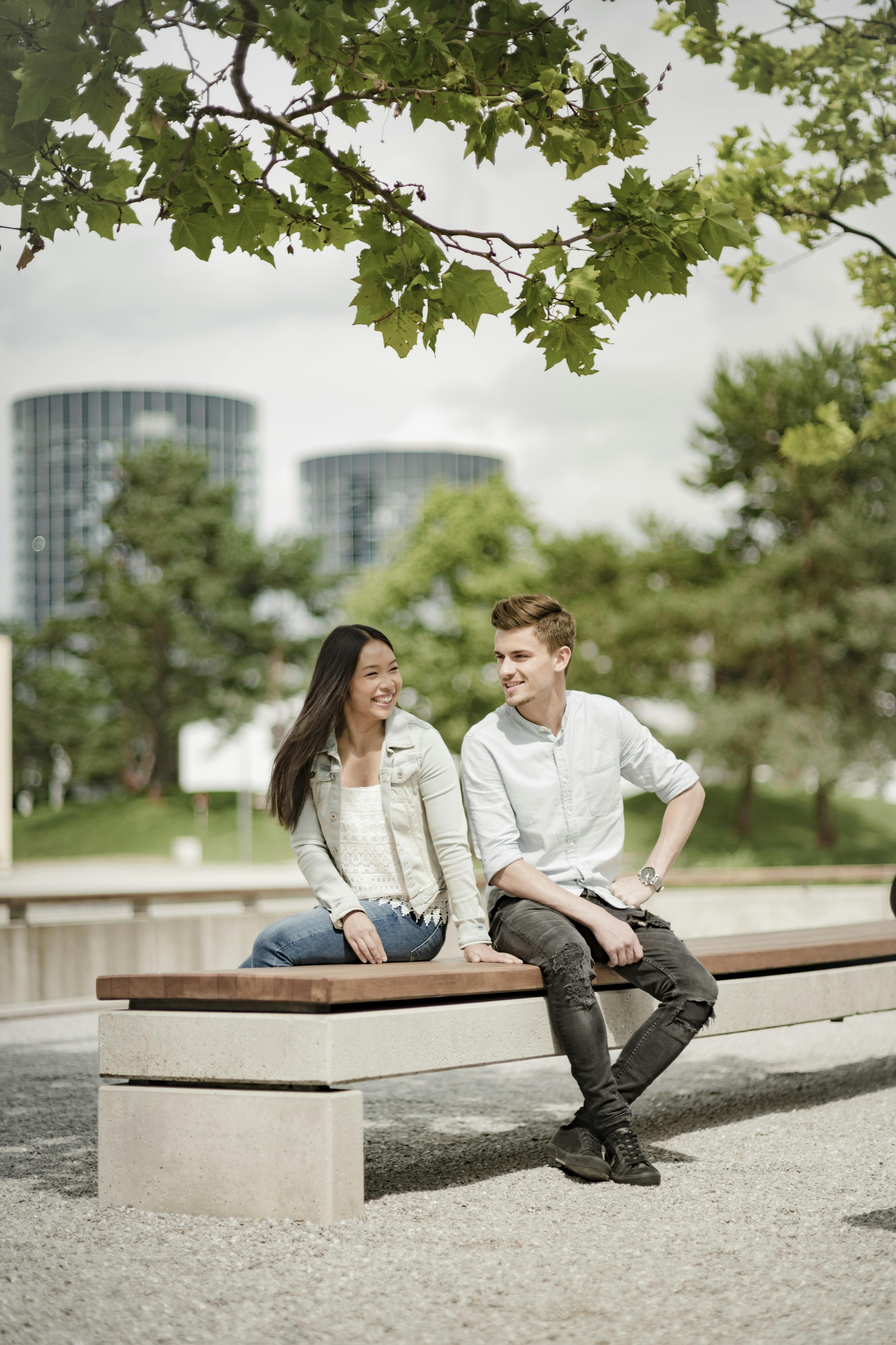 A smiling couple sits on a wooden bench in a park, with trees and two tall buildings in the background.
