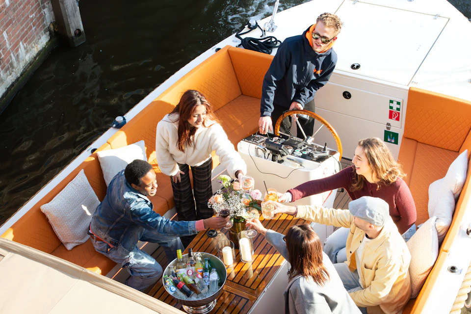 A group of people on a boat sharing drinks and smiling, with a man steering the boat in the background.