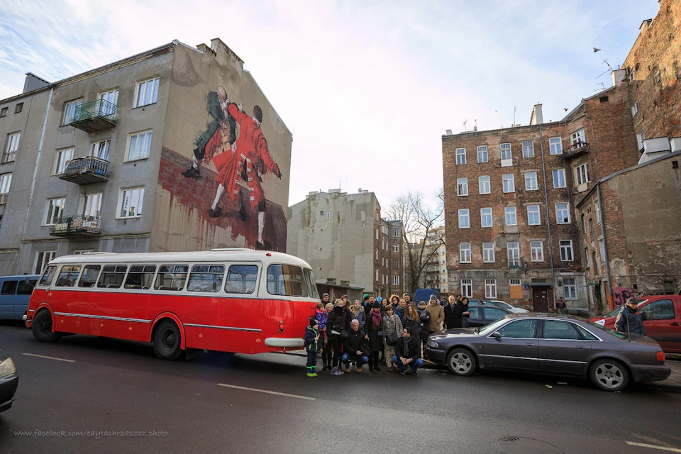 Group of people posing in front of a red and white bus, with buildings and a mural of dancing figures in the background.
