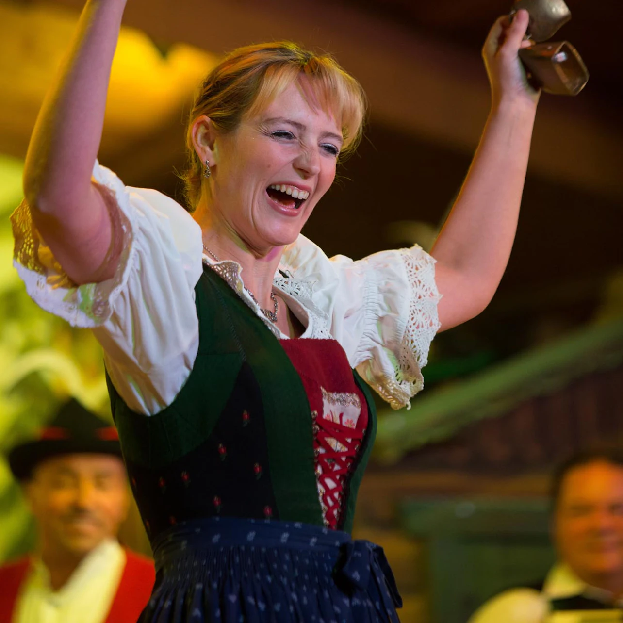 A woman in traditional Bavarian attire cheerfully lifts cowbells, with people smiling in the background.