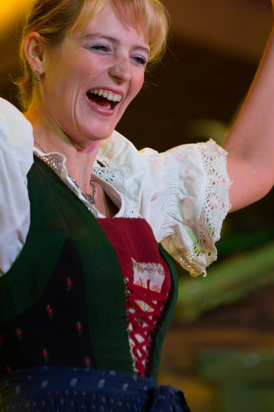 A woman in traditional Bavarian attire cheerfully lifts cowbells, with people smiling in the background.