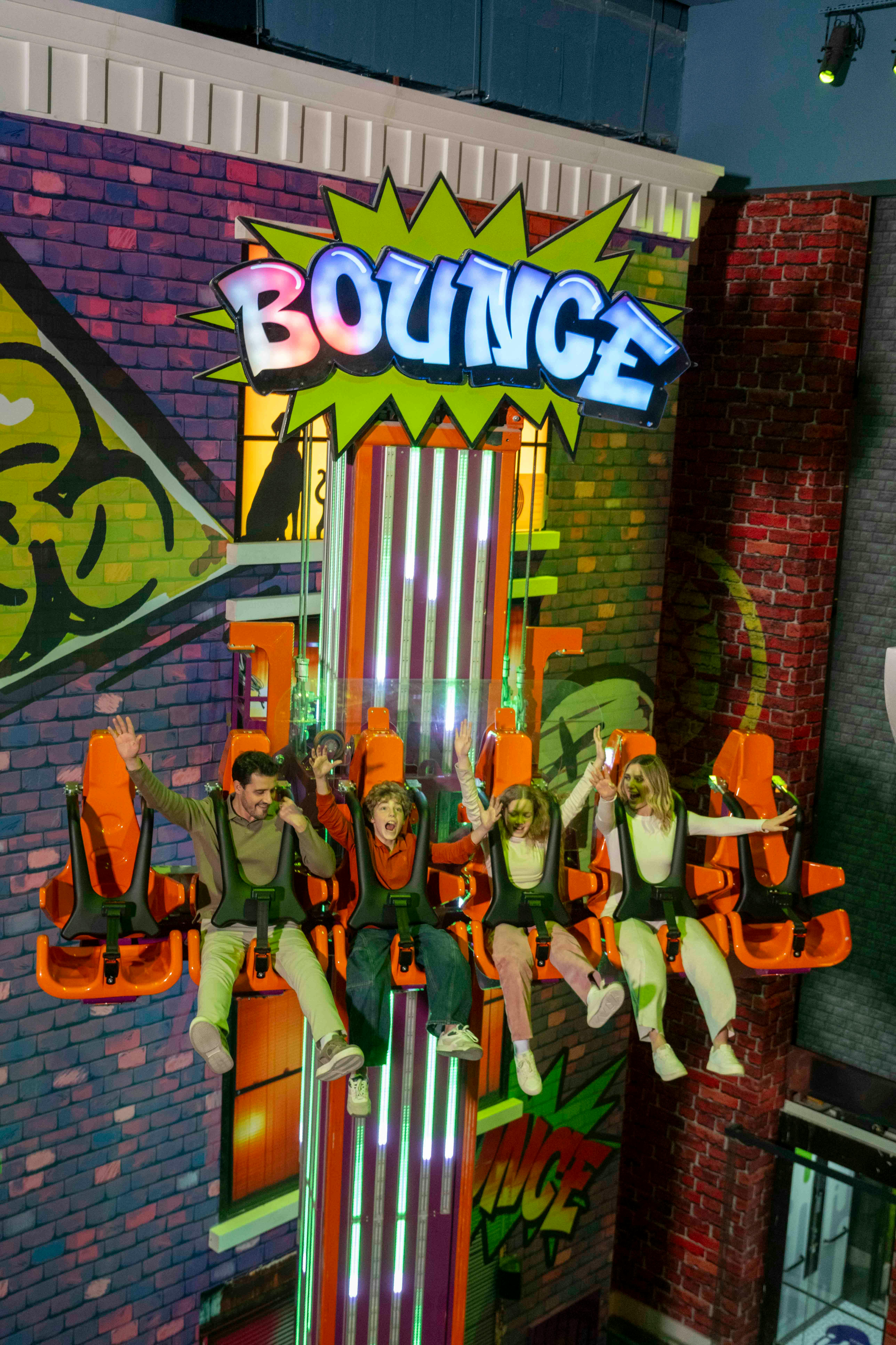 Vijf mensen zitten op een kleurrijke amusementsattractie en steken hun armen omhoog, met een achtergrond in comic-thema.