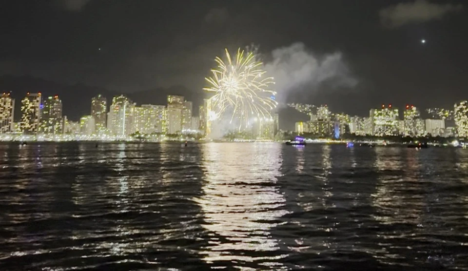 Fireworks illuminating the night sky above a city skyline, reflected in the water of a nearby harbor.