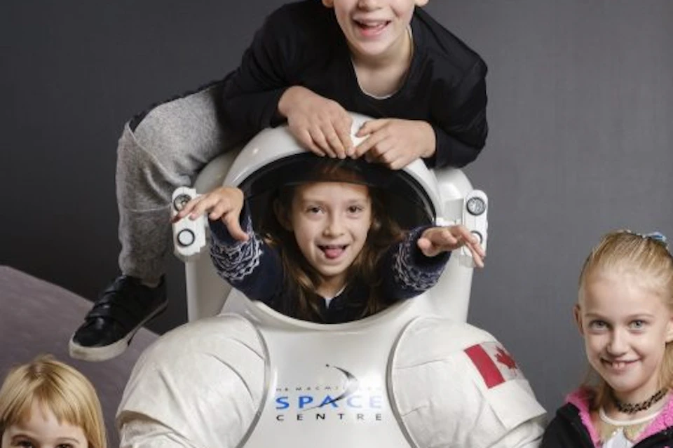 Four children posing with a space suit display; one child is inside the suit, another is climbing on top. Moon and Earth background.