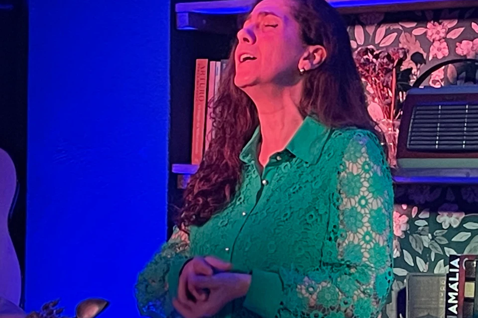 A woman sings passionately while two men play guitars beside her in a warmly lit room with floral wallpaper and shelves.