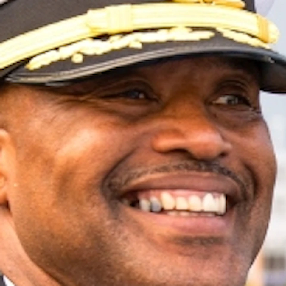 A man in a naval uniform smiles while standing on a ship’s deck with a waterfront cityscape in the background.