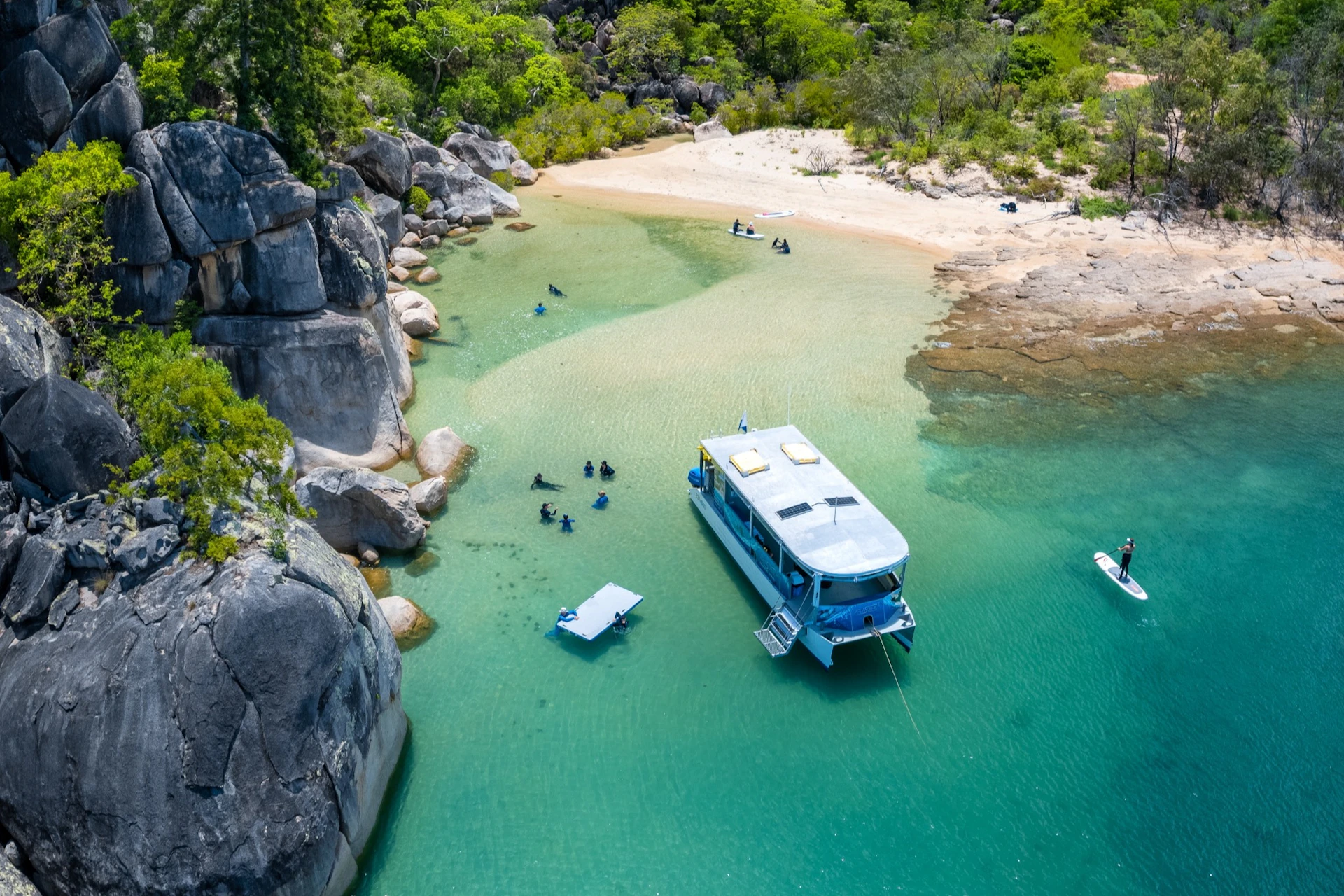 A boat anchored in clear turquoise water near a rocky shoreline and a small sandy beach, with people swimming and exploring.