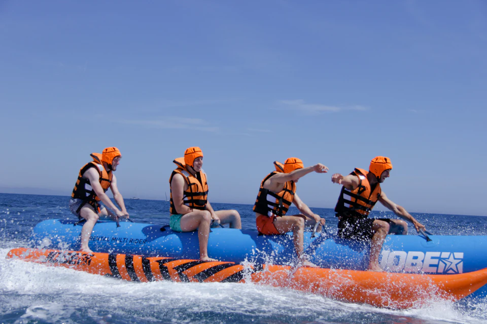 Four people wearing orange life vests and helmets ride an orange and blue inflatable banana boat on the sea under a clear blue sky.