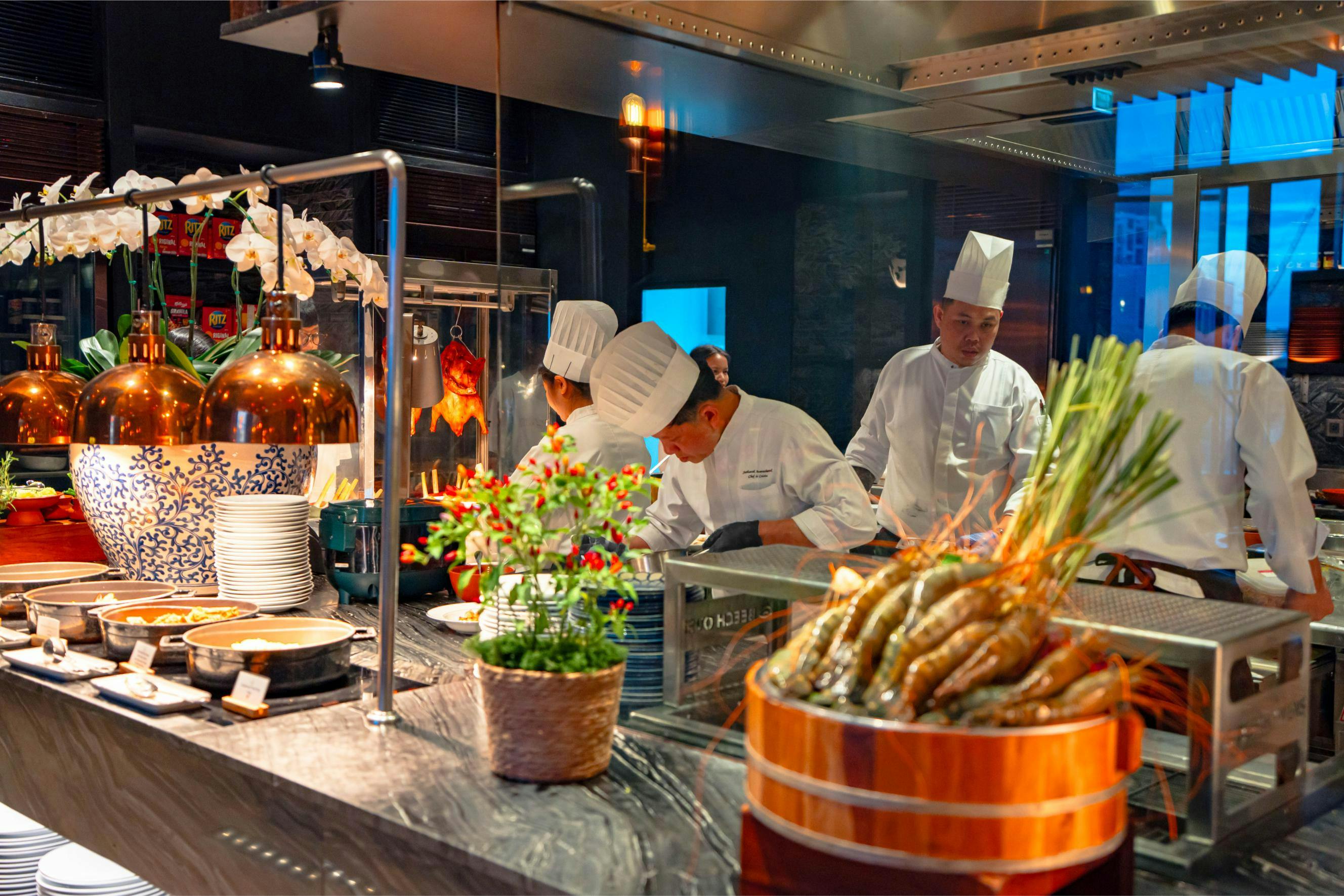 Chefs in white uniforms and hats working in a modern kitchen with fresh ingredients and cooking equipment visible.