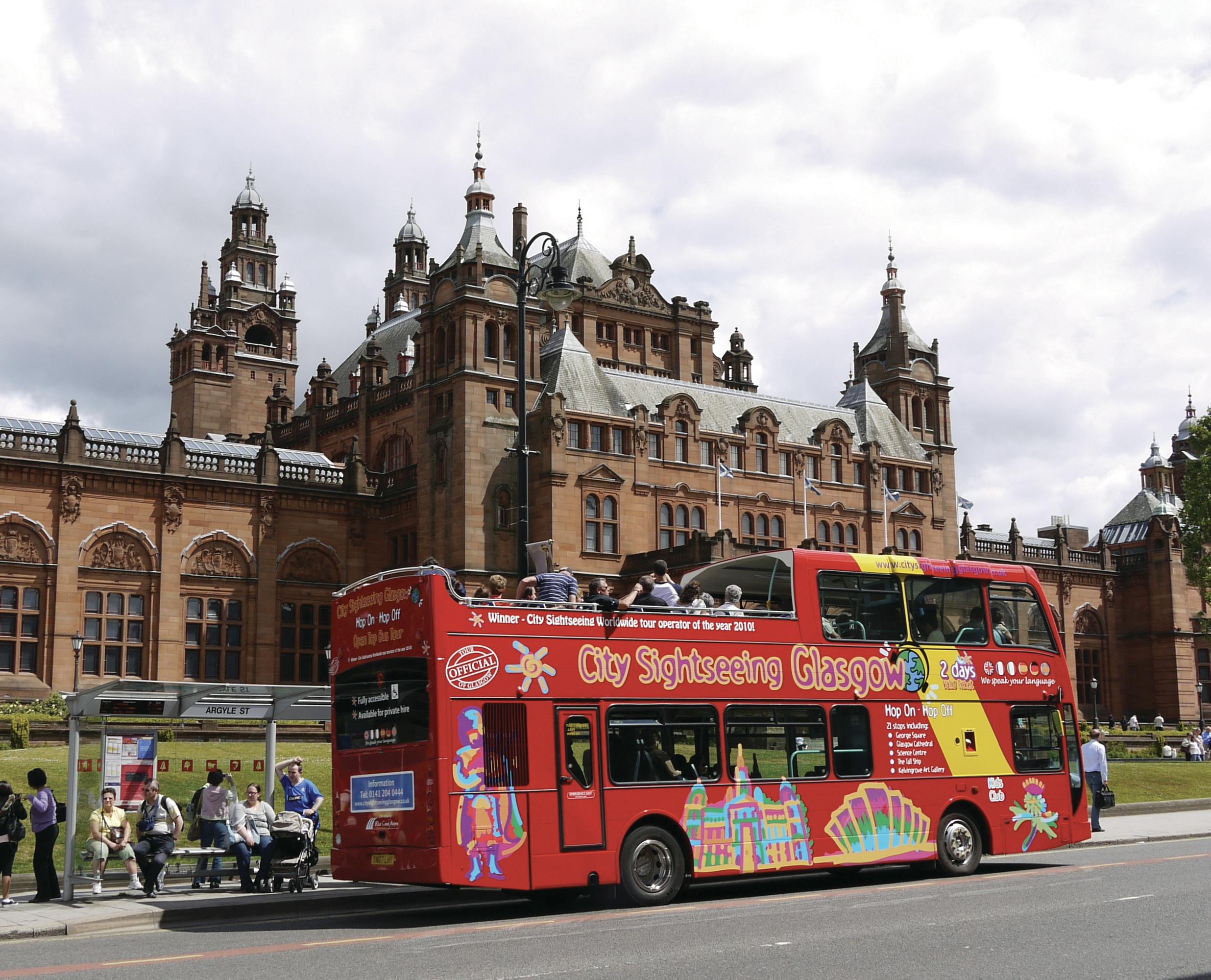 Ένα κόκκινο διώροφο τουριστικό λεωφορείο με το &#34;City Sightseeing Glasgow&#34; σταθμευμένο μπροστά από ένα ιστορικό, περίτεχνο κτίριο.