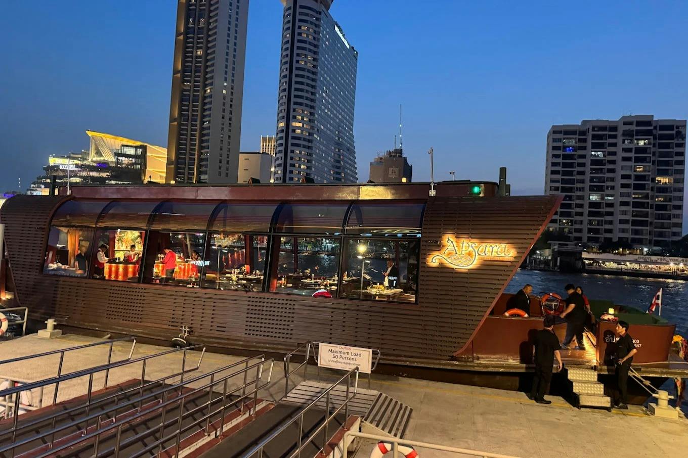 A docked boat named "Apsara" illuminated at night, with a cityscape and tall buildings in the background.