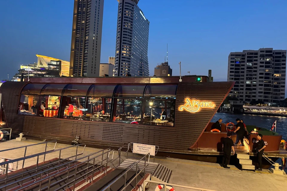 A docked boat named "Apsara" illuminated at night, with a cityscape and tall buildings in the background.