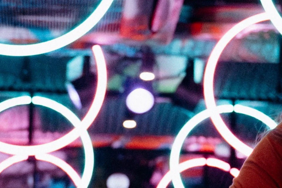 A woman energetically riding a stationary bike in a colorful, neon-lit studio with circular light designs on the walls.