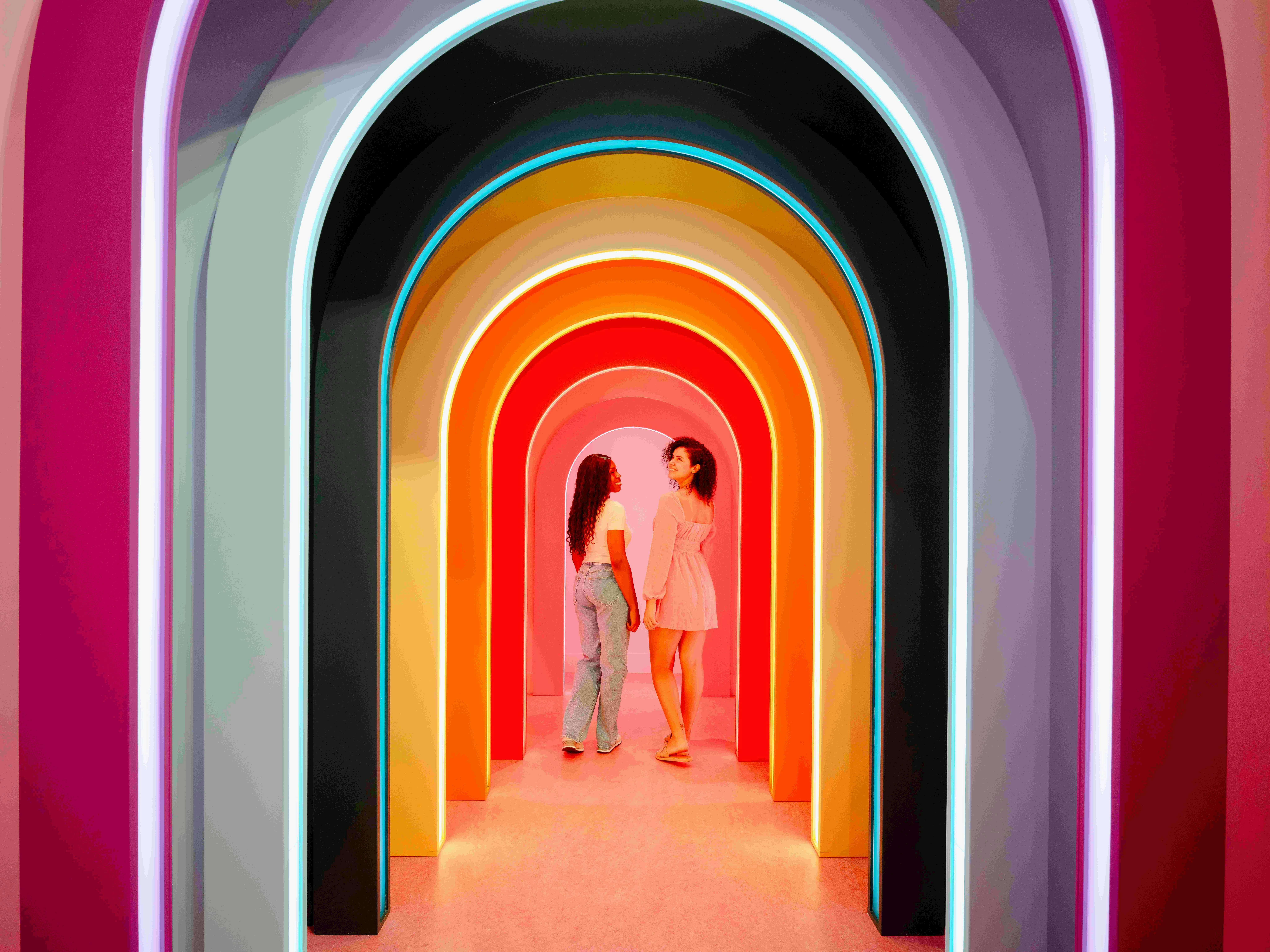 Dvě ženy stojící pod řadou barevných, osvětlených oblouků, čelem k sobě a usmívající se na sebe.