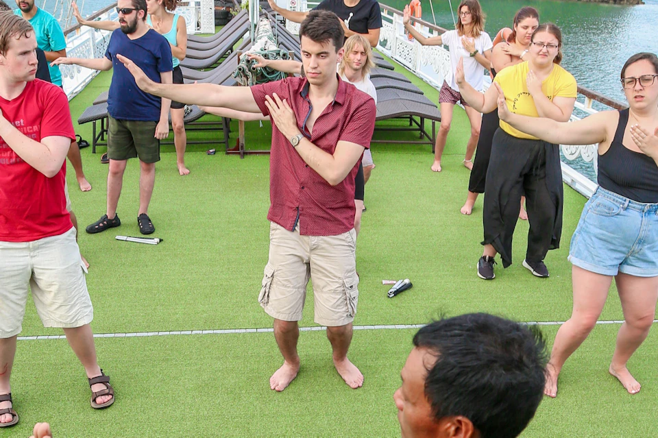 A group of people practicing Tai Chi on a boat deck, led by an instructor wearing a red robe, with rocky cliffs in the background.