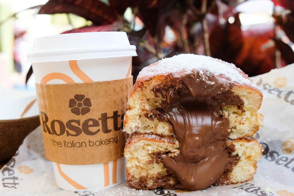 A cream-filled pastry dripping with chocolate alongside a coffee cup with a brown sleeve labeled "Rosetta the Italian bakery."