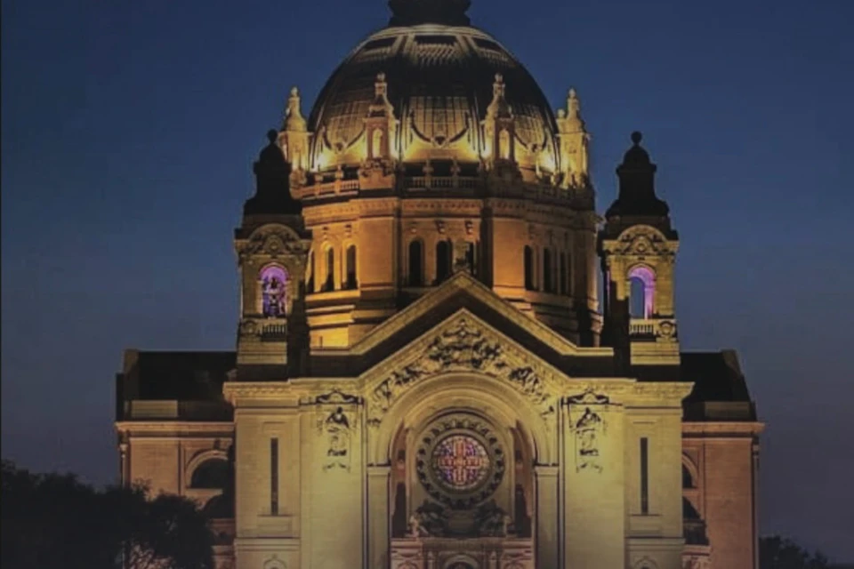 A lit-up cathedral dome and facade at night, featuring intricate architectural details and stained glass windows.