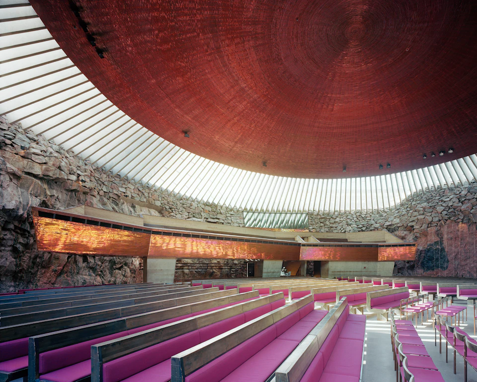 A spacious room with a circular, wood-textured ceiling, glass walls, rock formations, and rows of purple-cushioned pews.