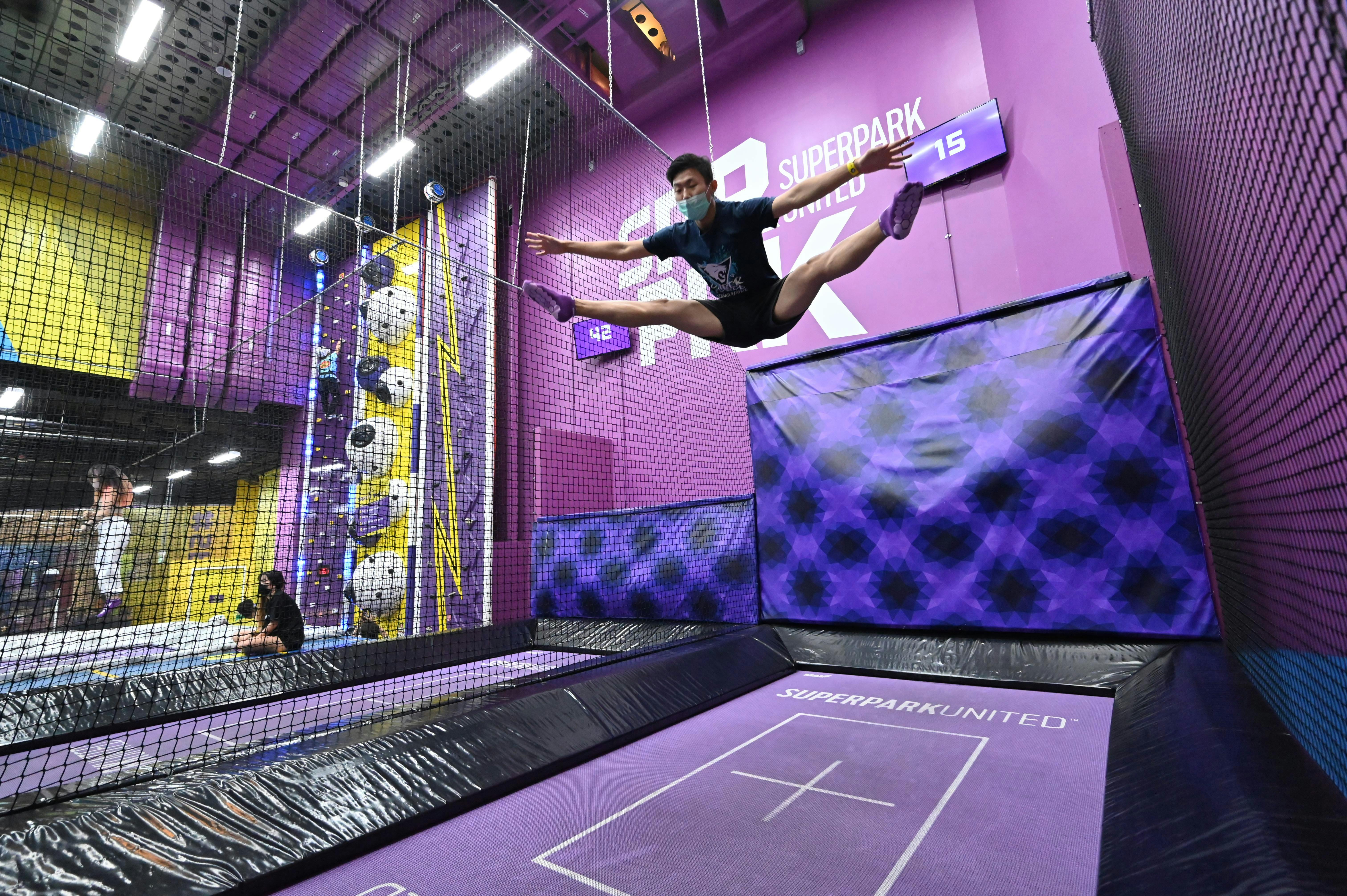 Person in mid-air split jump on trampoline in purple indoor park, with climbing wall and netting in the background.