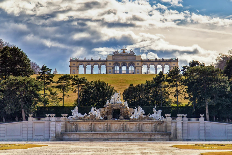 A grand stone fountain with sculptures in front of a hill, topped by a large colonnaded structure under a cloudy sky.