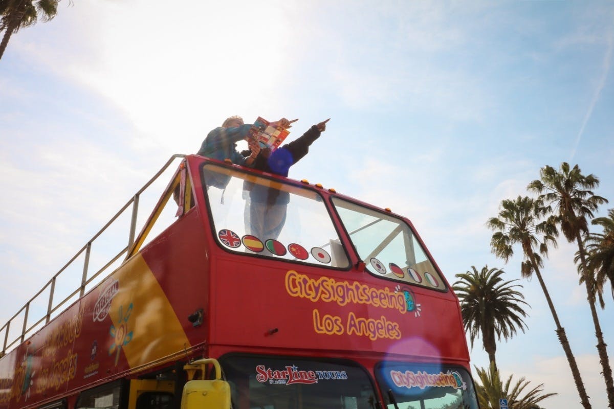 Two people stand on the open top of a red CitySightseeing tour bus, holding a map and pointing, with palm trees in the background.