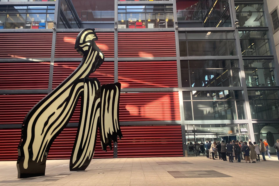 A modern building with red and glass panels, featuring a large abstract sculpture in the foreground.