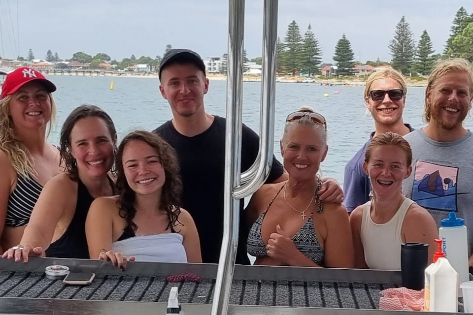 A group of smiling people on a boat, with a waterfront and trees visible in the background. A sign reads "EMERGENCY ONLY."