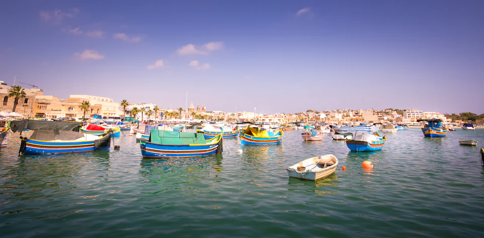 A harbor with colorful wooden boats floating on calm water under a clear, bright sky; town buildings visible in the distance.