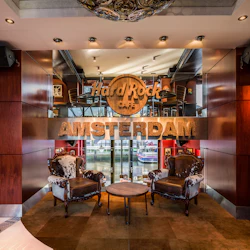 Två vintage stolar och ett runt bord framför en "Hard Rock Cafe Amsterdam" -skylt, med glasdörrar som visar utsikt över vattnet.