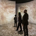 Three people stand in a room with large, curved projections of a detailed, textured image, possibly artwork, covering the walls and floor.