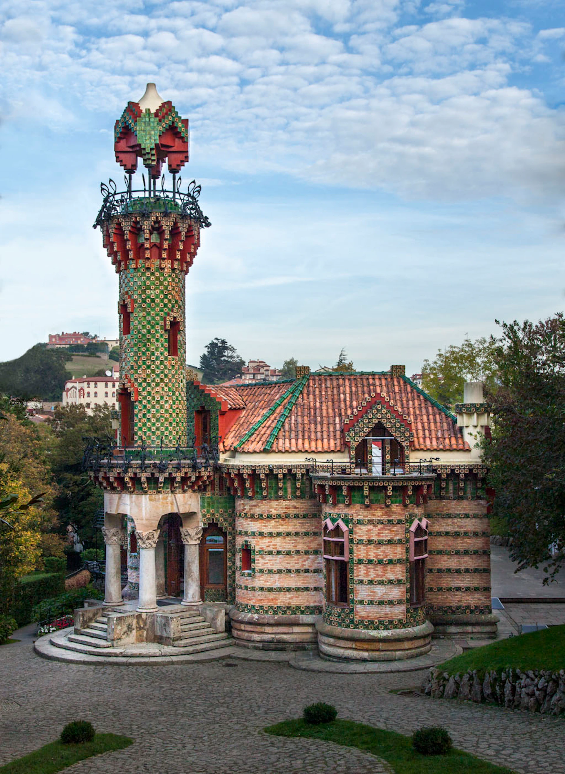 A decorative building with a red-tiled roof and a mosaic-covered tower, surrounded by greenery on a sunny day.