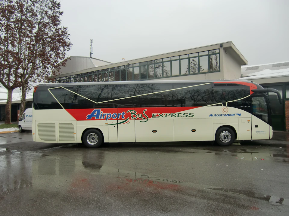 An "Airport Bus Express" parked beside a building on a wet road under a cloudy sky.