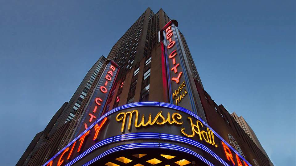 Tall building with bright neon signs reading "Radio City Music Hall" against a twilight sky.