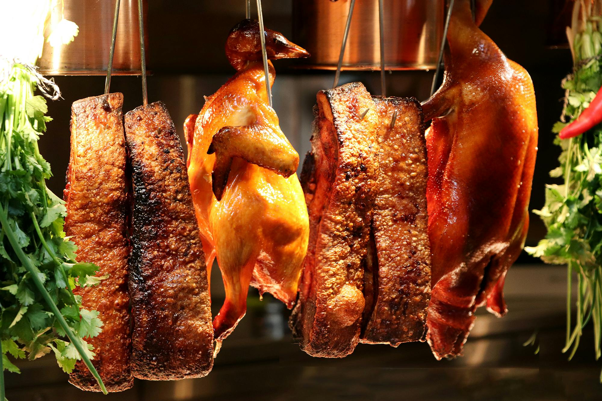 Roasted meats, including duck and pork, hanging on hooks in a food display under warm lighting.