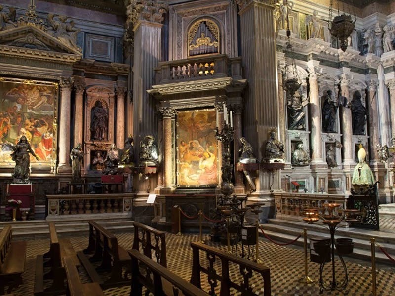Interno di una chiesa decorata con dipinti religiosi, statue, banchi in legno e colonne dettagliate, con illuminazione soffusa.