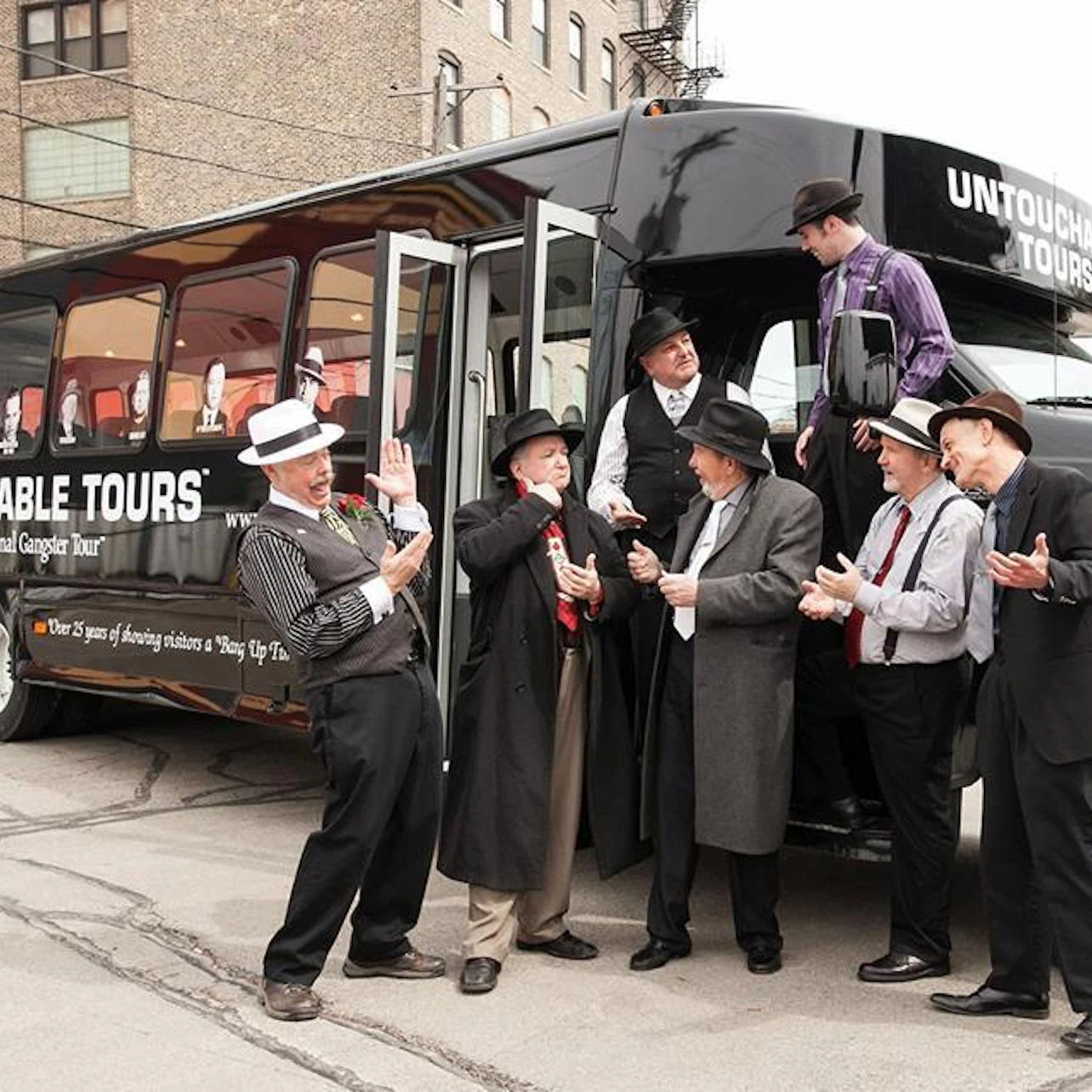 A group of men in retro-style outfits posing near a black tour bus labeled "Untouchable Tours" in an urban setting.