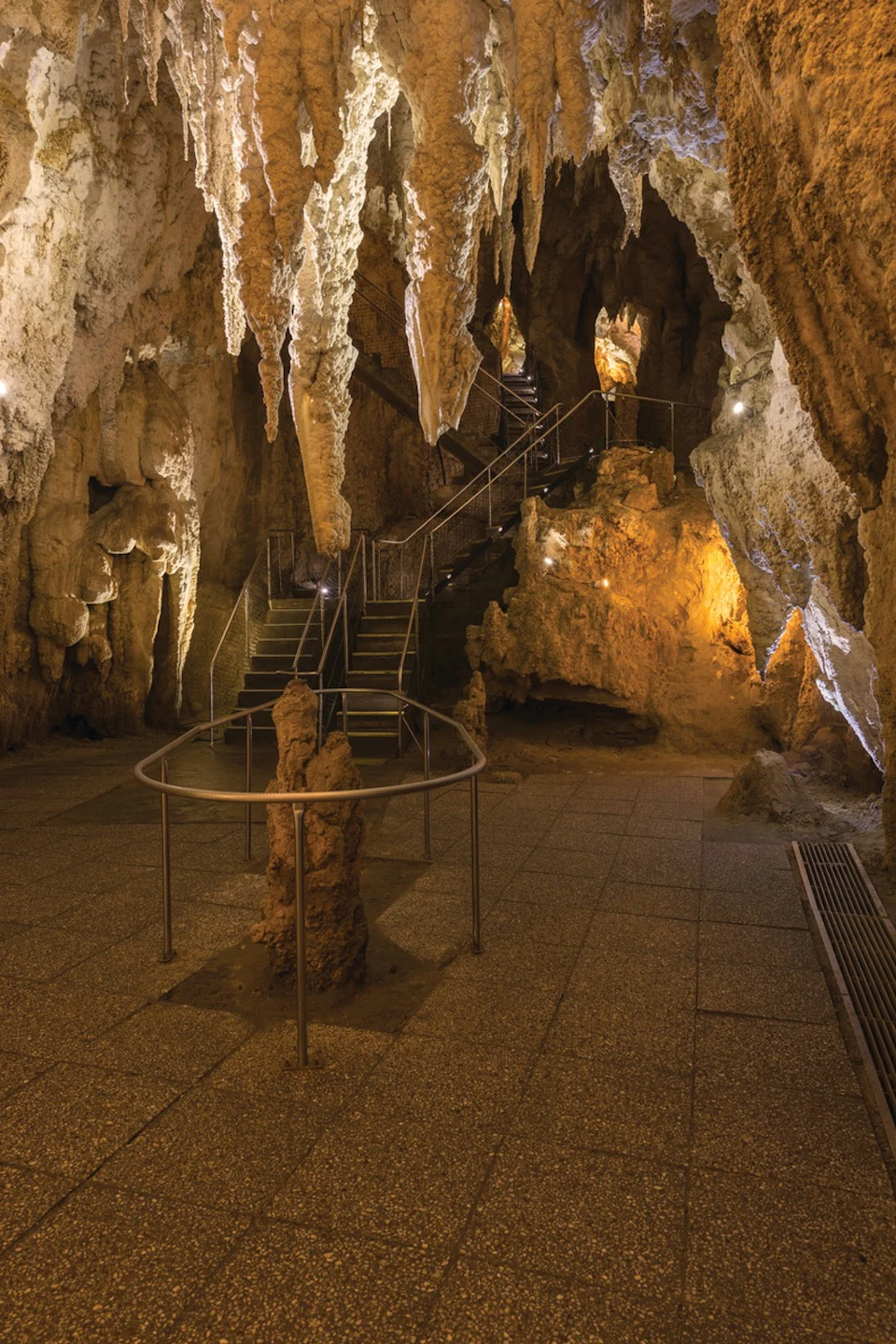 Interior of a cave with stalactites and stalagmites, a staircase with railings, and pathway lighting.