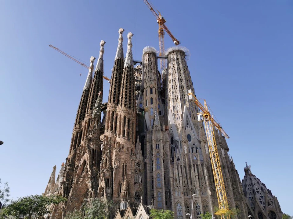 A large, detailed church structure with multiple tall spires under construction, featuring several cranes, against a clear blue sky.