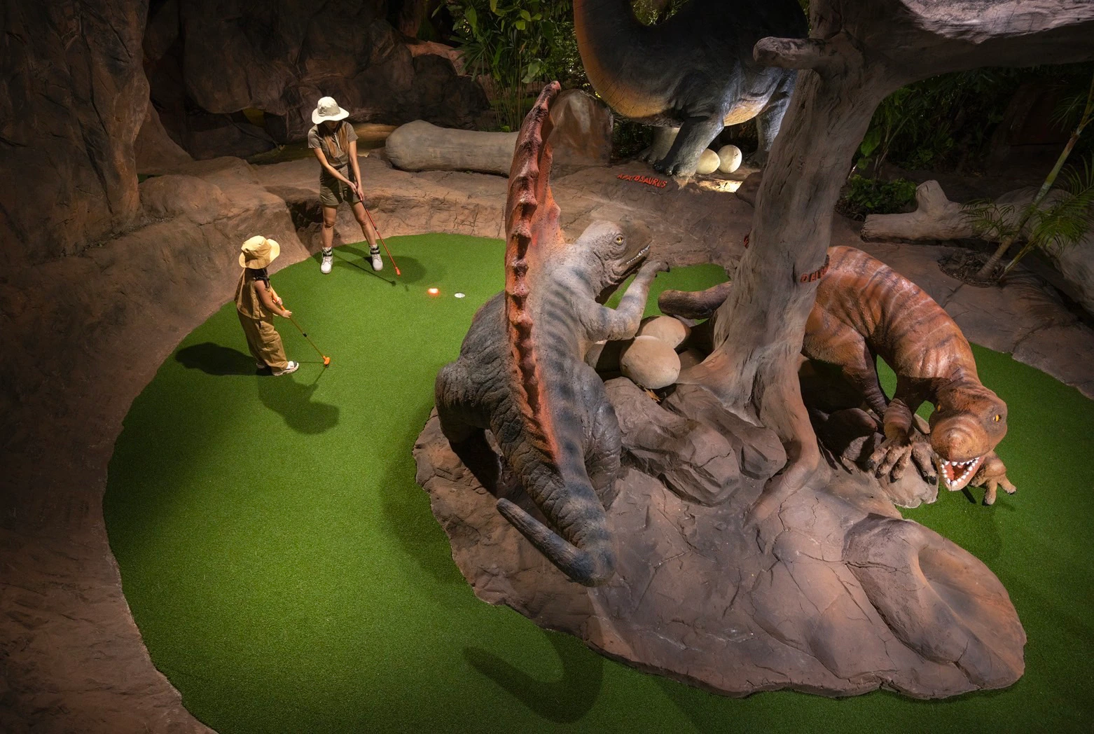 Two children play mini-golf on a course featuring dinosaur statues and artificial rocks in a jungle-themed setting.