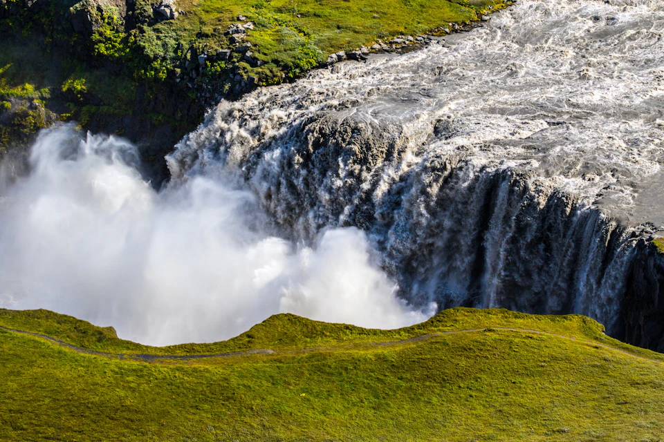 A powerful waterfall cascades into a misty pool, surrounded by lush green grass and rocky terrain.