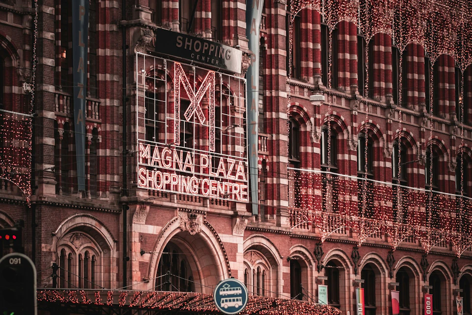 Brick facade of Magna Plaza Shopping Centre with light decorations and a bus stop sign in front.