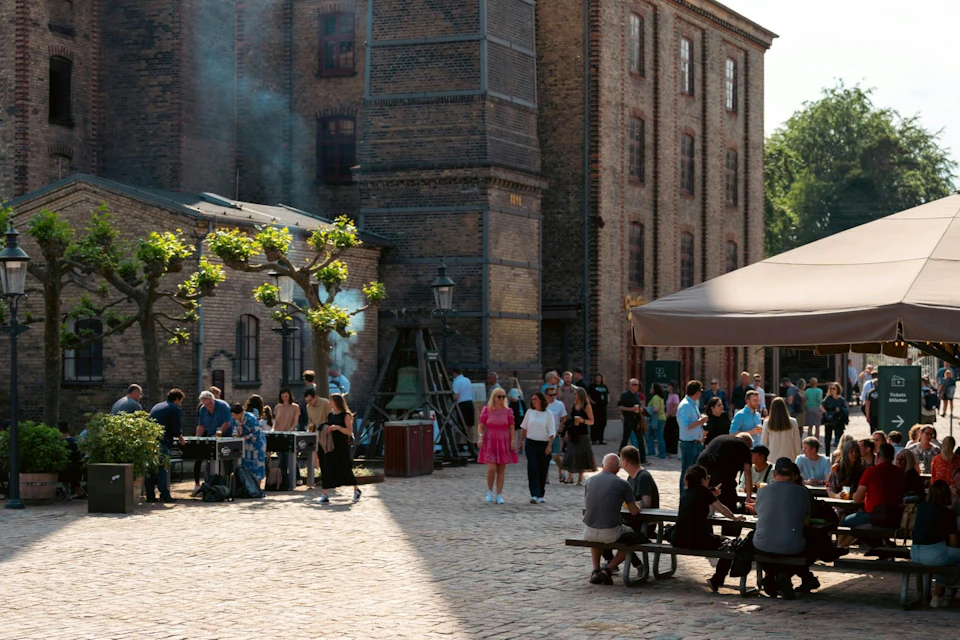 People gathering in an outdoor courtyard near brick buildings, some seated at tables, others standing under a canopy.