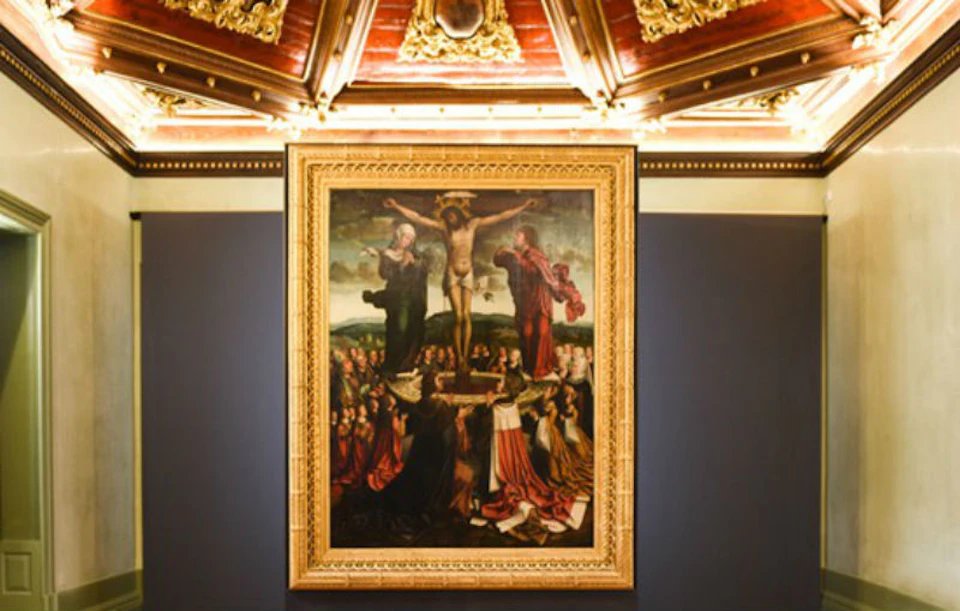 A framed painting of the crucifixion of Jesus, surrounded by mourners, mounted on a wall with an ornate ceiling above.