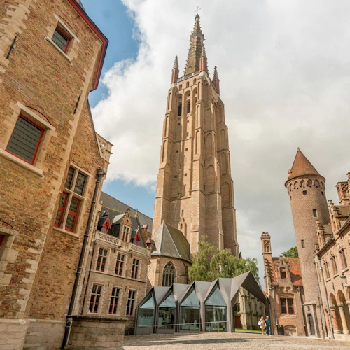 Ett högt, historiskt tegeltorn omgivet av gamla byggnader med välvda fönster och röda accenter under en delvis molnig himmel.