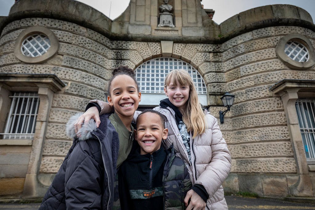 Three children, two girls and one boy, smile and pose together in front of a historic stone building with barred windows.